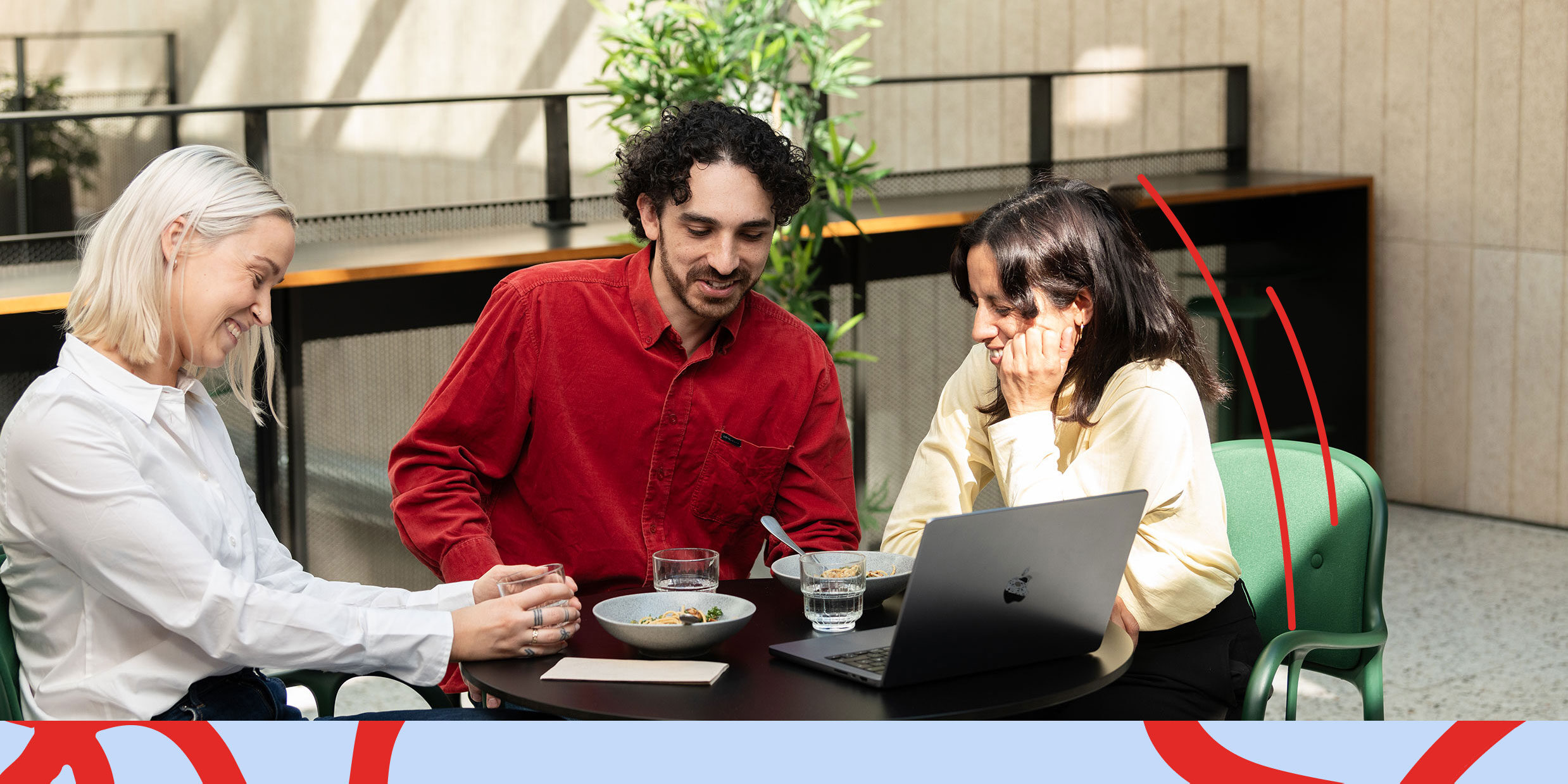 Three colleagues sitting at a table with a laptop, laughing and engaged in discussion, in a bright indoor space with plants.