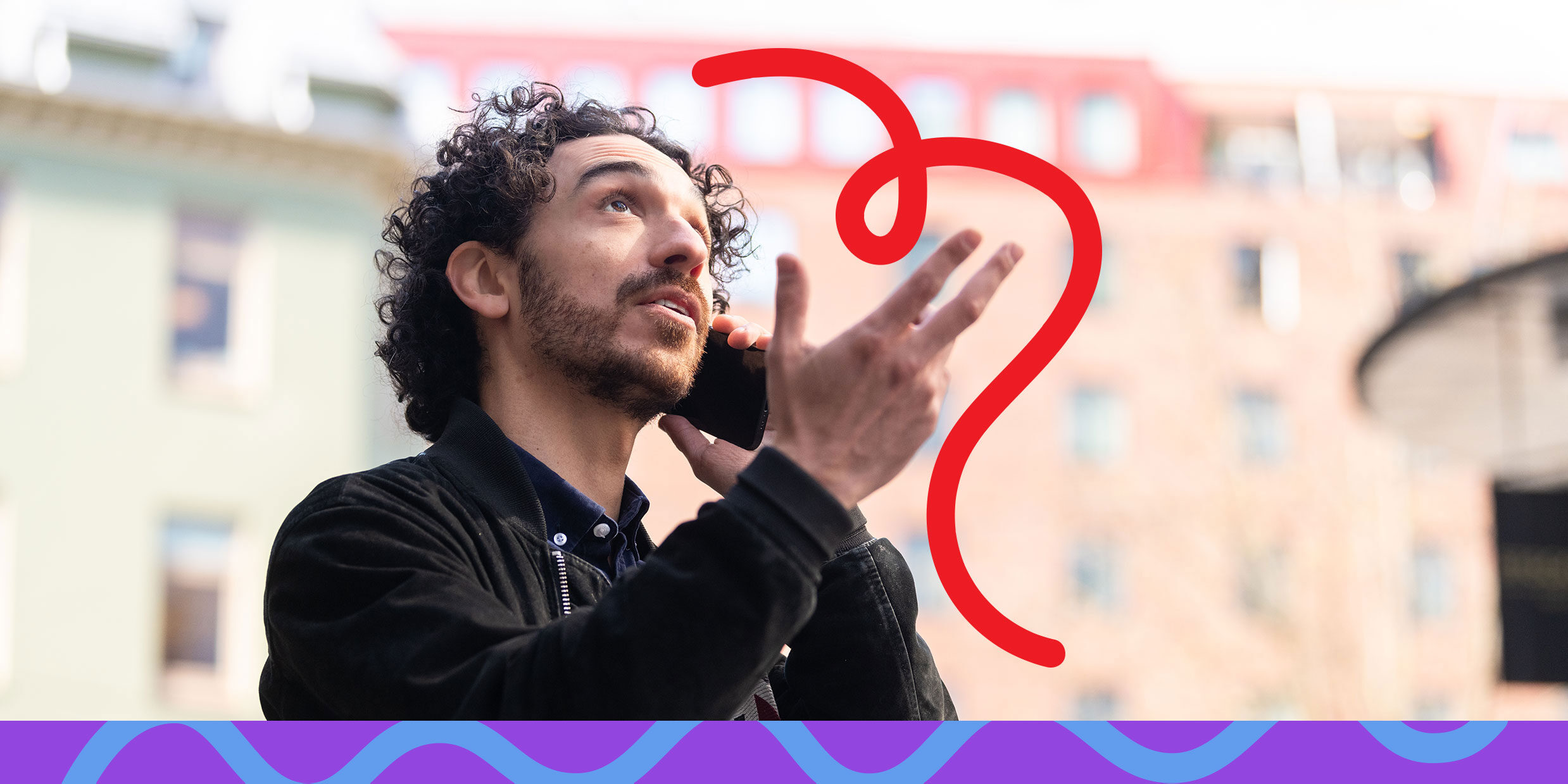 A man with curly hair talking on his phone outside, gesturing with his hand, with buildings in the background.